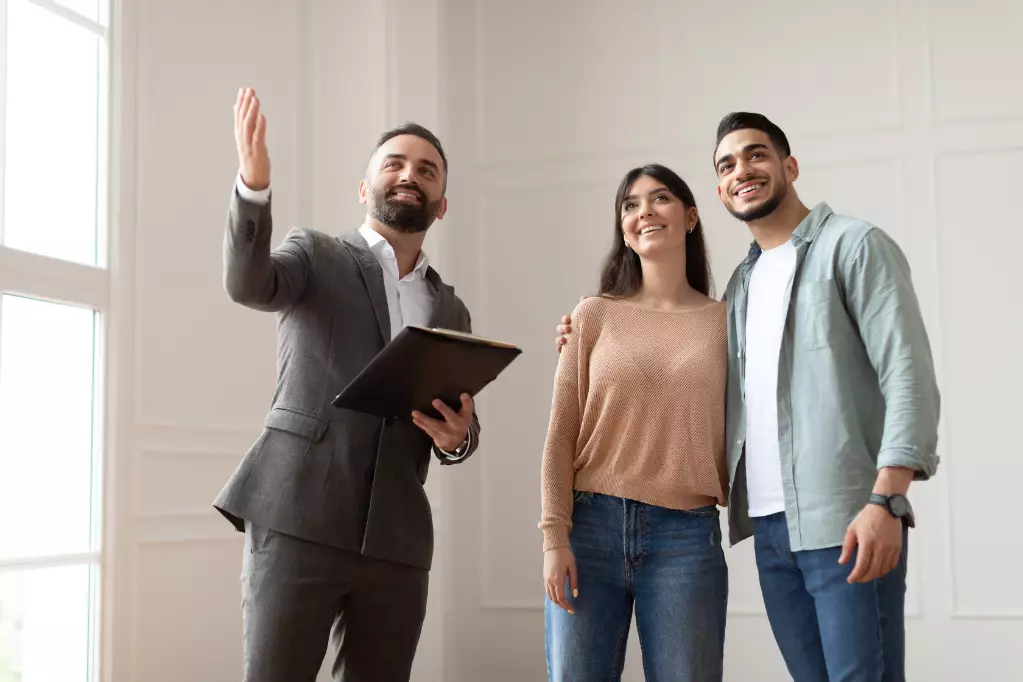 Portrait Of Male Realtor Or Architect Wearing Suit Showing New Empty House To Smiling Millennial Buyers, Pointing. Husband Embracing His Wife, Visiting Residential Building, Choosing Home For Family